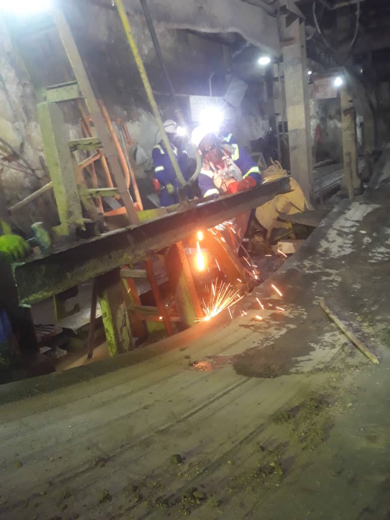 56168147-IMG-20230509-WA0028 Workers welding metal structures underground, performing engineering solutions for mining operations in a dimly lit industrial setting.